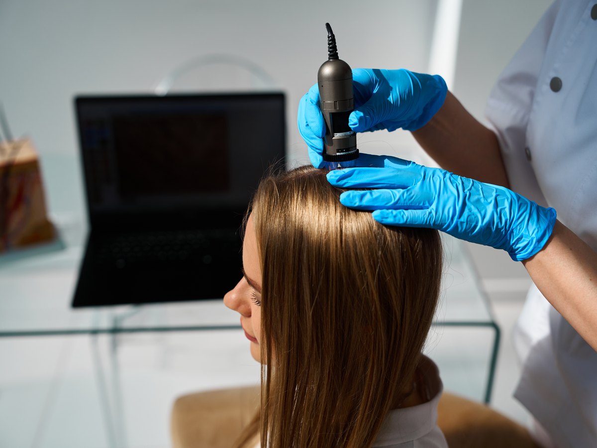 Especialista con guantes examinando con un dermatoscopio digital el cuero cabelludo de una paciente en consulta