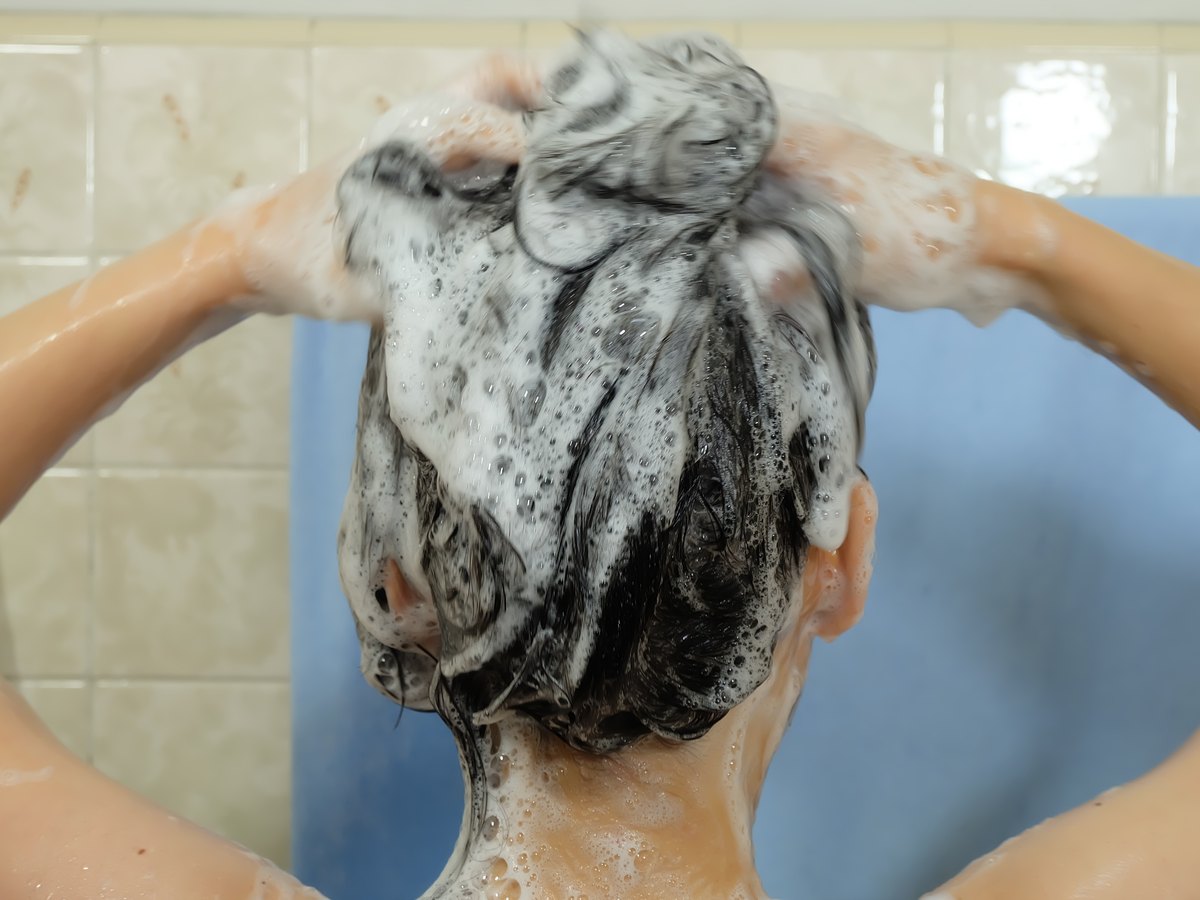 Mujer lavándose el pelo en la ducha con abundante espuma de champú, vista de espaldas