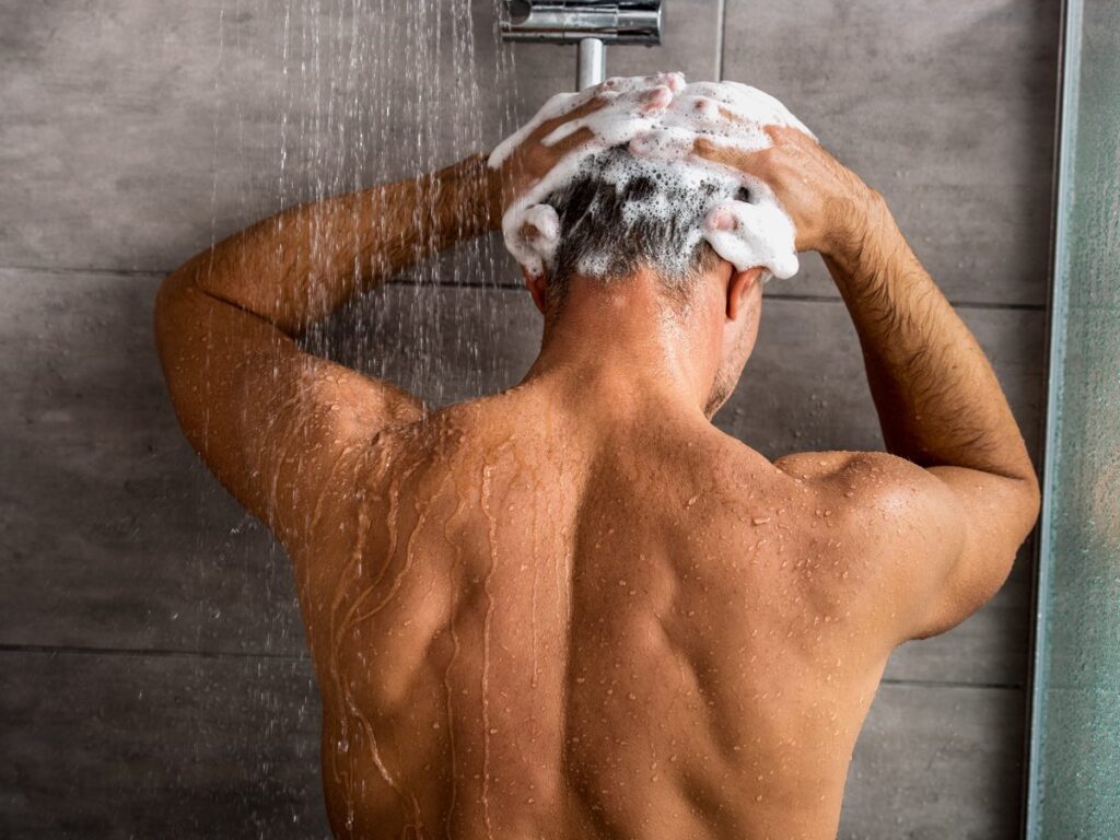 Hombre de espaldas lavándose el pelo con champú bajo la ducha