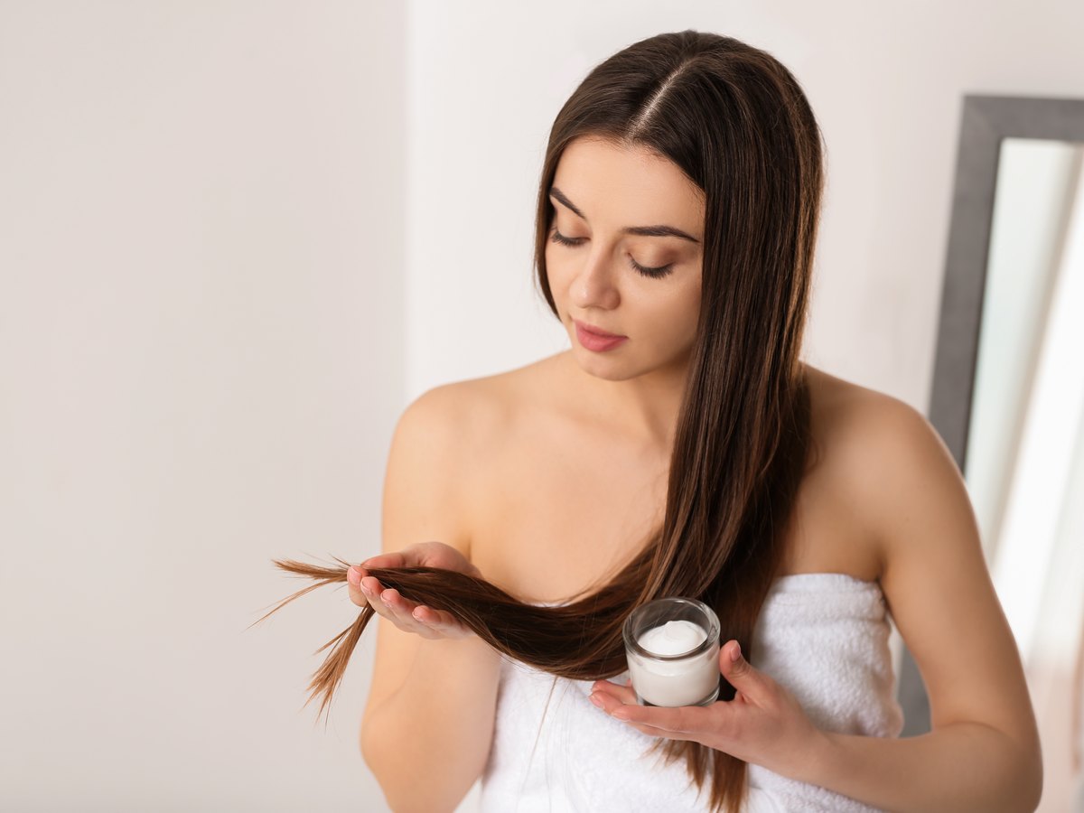 Mujer envuelta en una toalla con un tarro de mascarilla capilar en la mano, revisándose las puntas del pelo largo