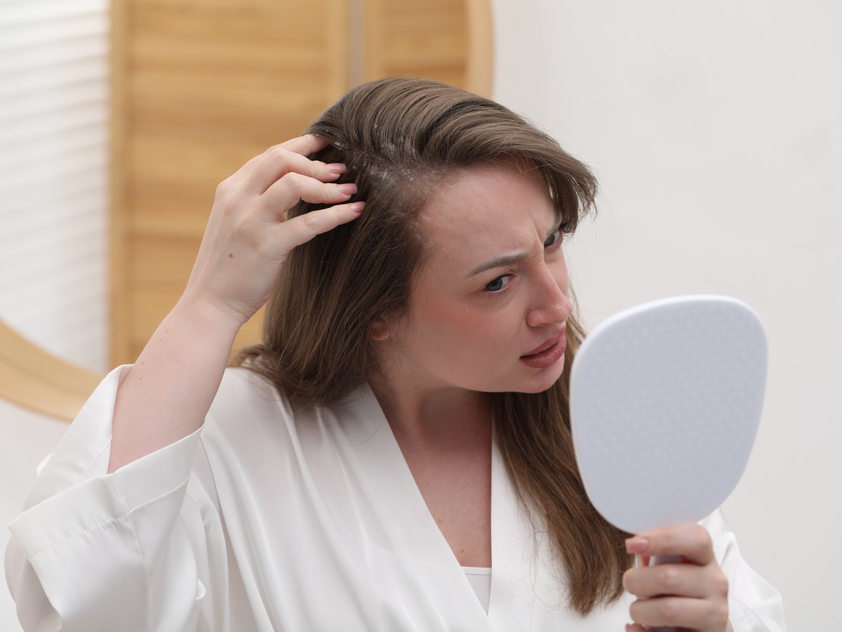 Mujer apartándose el pelo y mirándose la raya con un espejo de mano en el baño